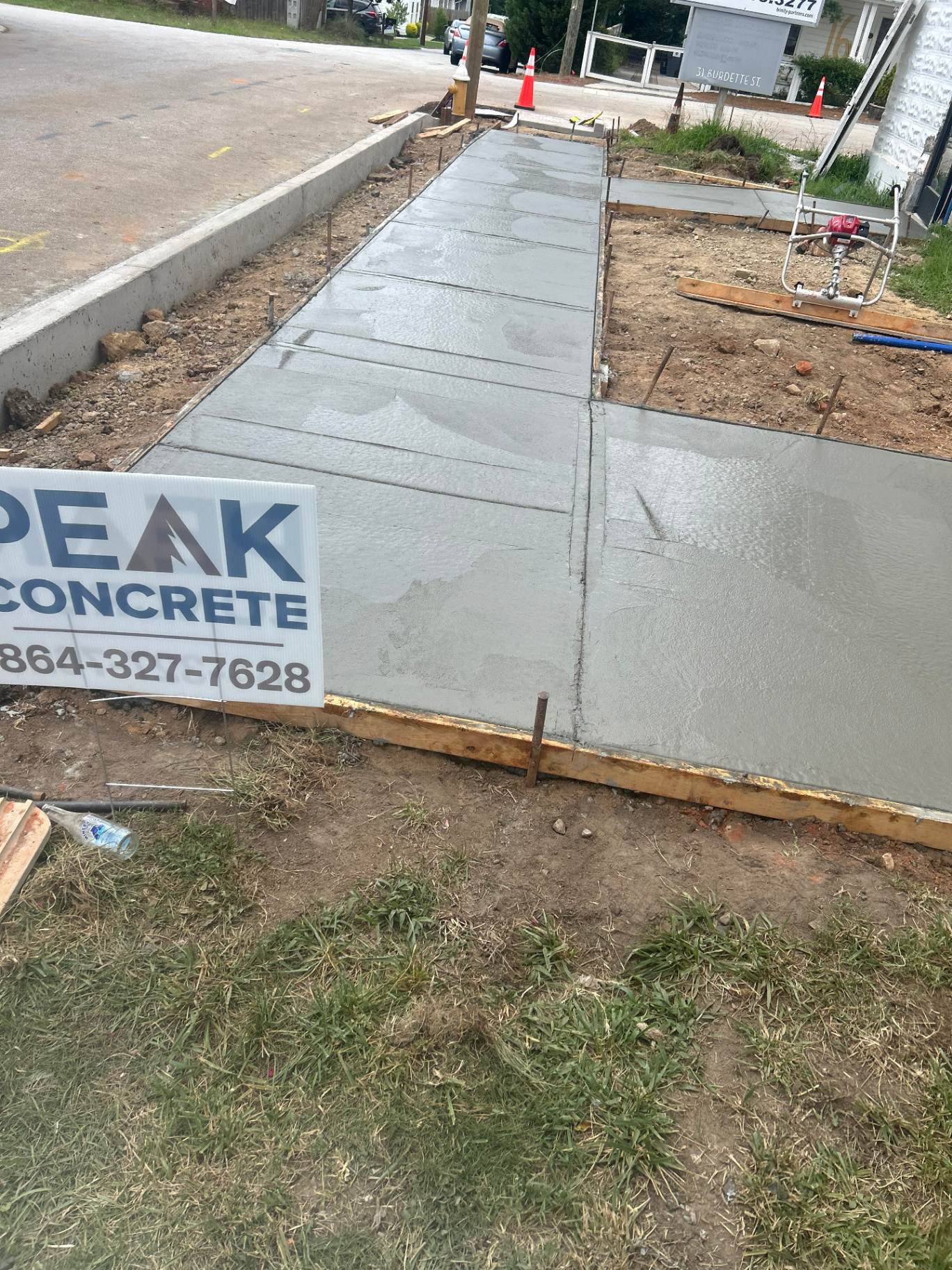 Completed section of broom-finished concrete sidewalk with control joints alongside a new curb, marked with a Peak Concrete yard sign.
