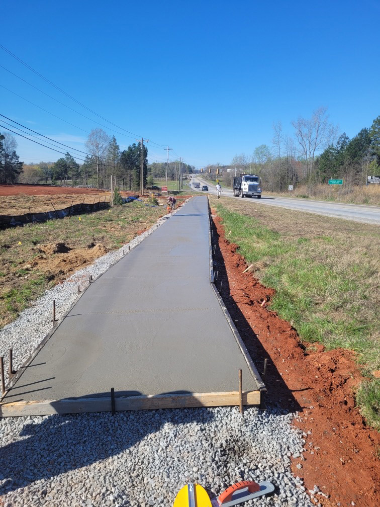 Long freshly poured roadside sidewalk still wet in the forms alongside a state highway, with a dump truck passing in the background.