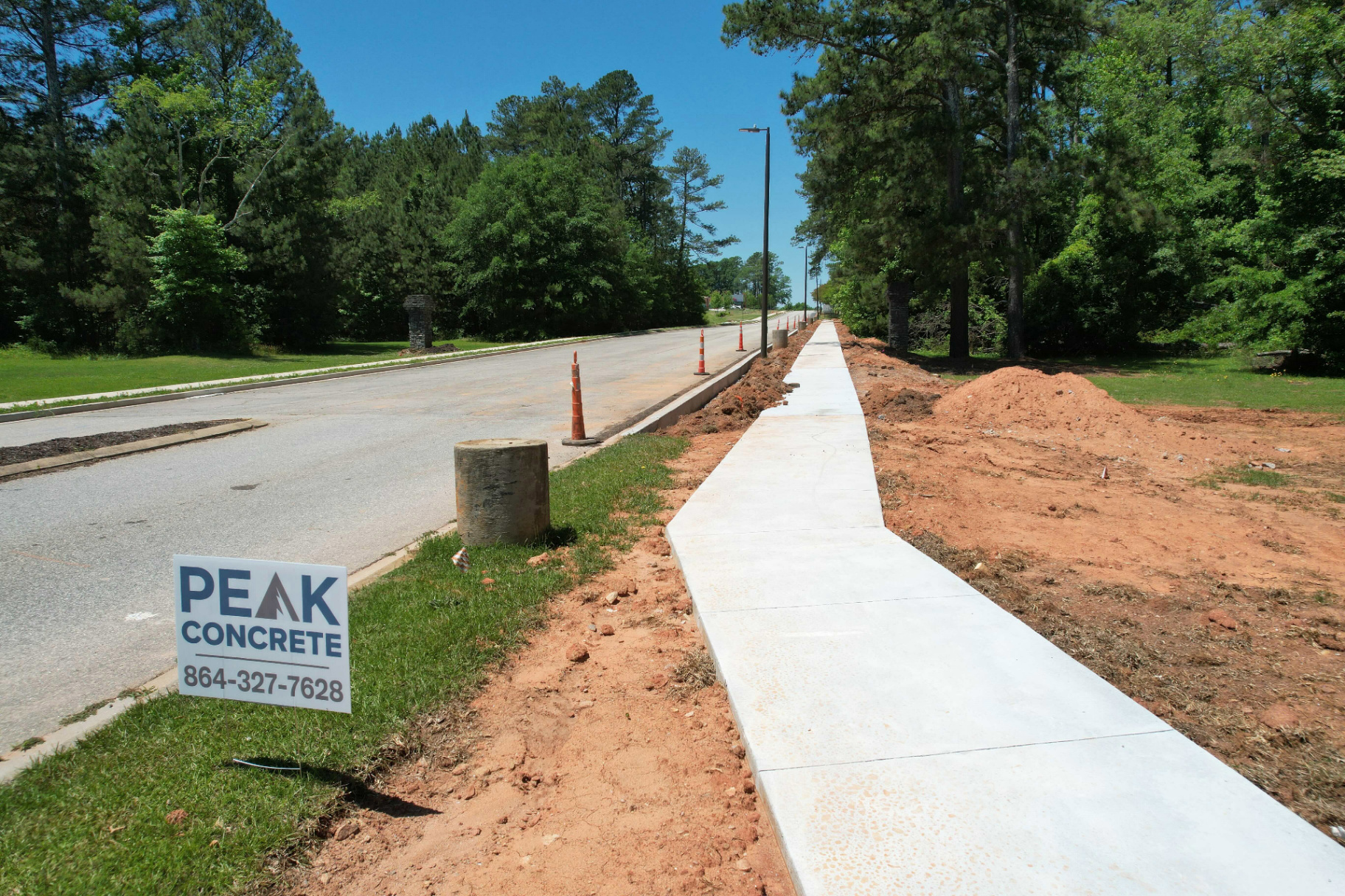 Newly poured concrete curb and sidewalk running alongside a roadway with a Peak Concrete yard sign in the foreground, red clay backfill still exposed.