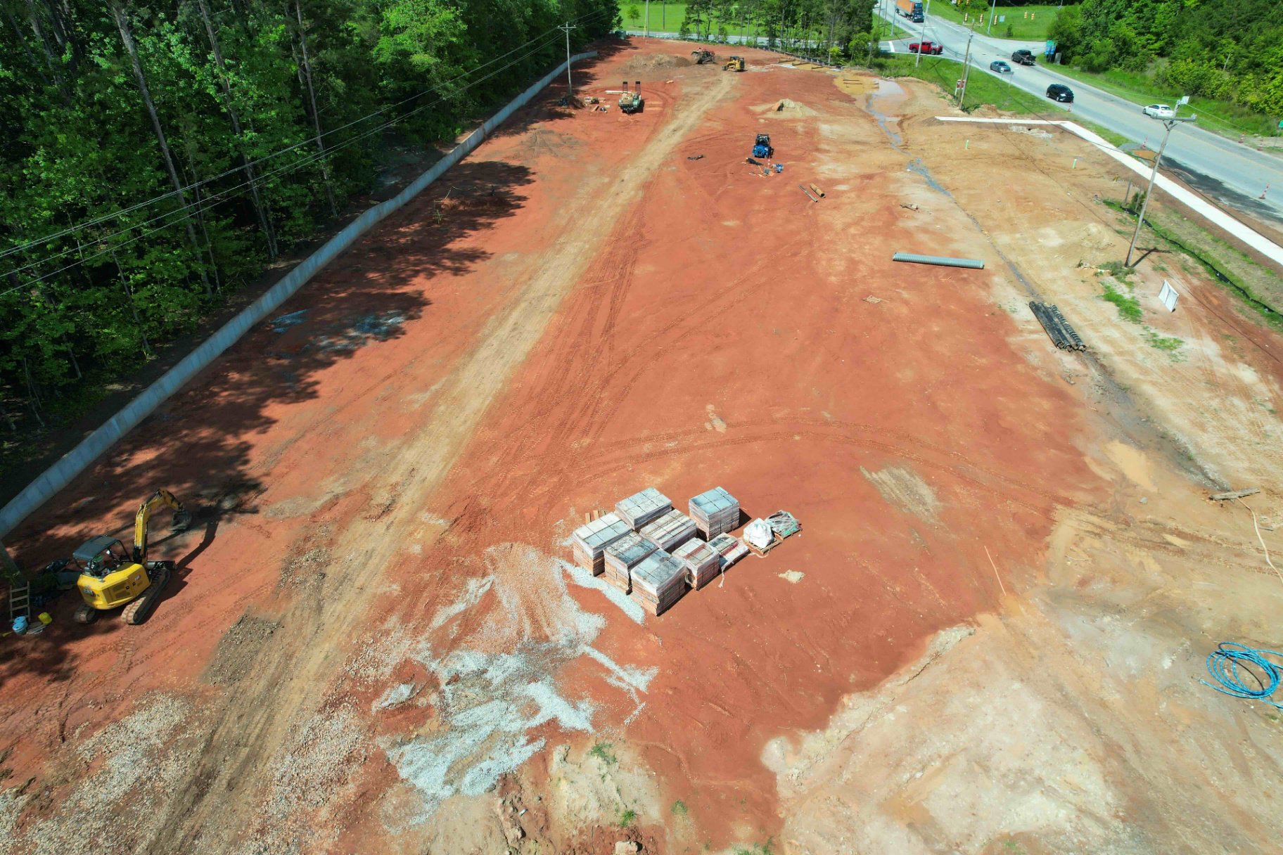 Aerial view of a graded commercial site with a long finished concrete retaining wall along the tree line, red clay pad ready for building construction, pallets of block staged on site.
