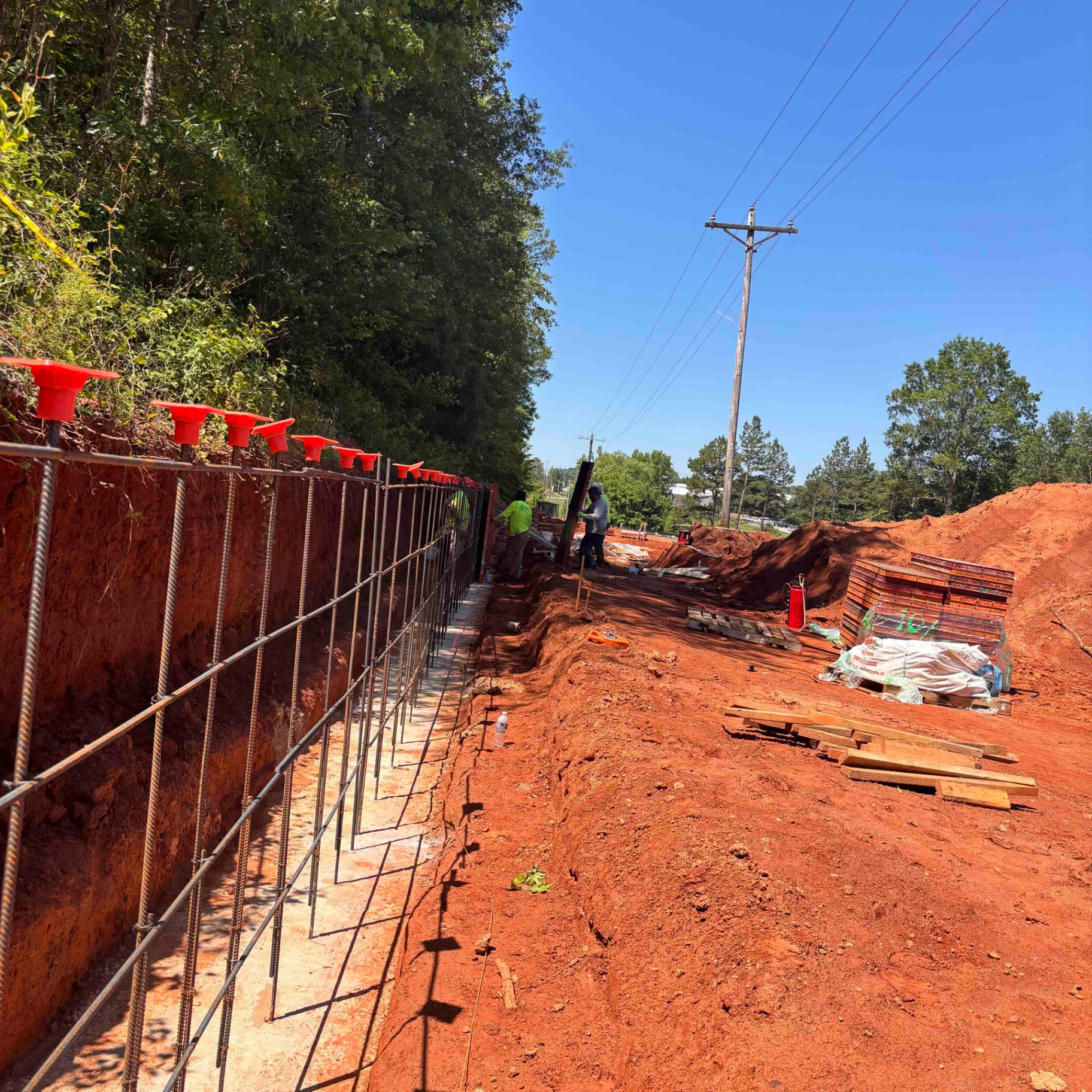 Cast-in-place concrete retaining wall footing with vertical rebar cage and orange rebar safety caps, crew setting formwork against a red clay embankment.