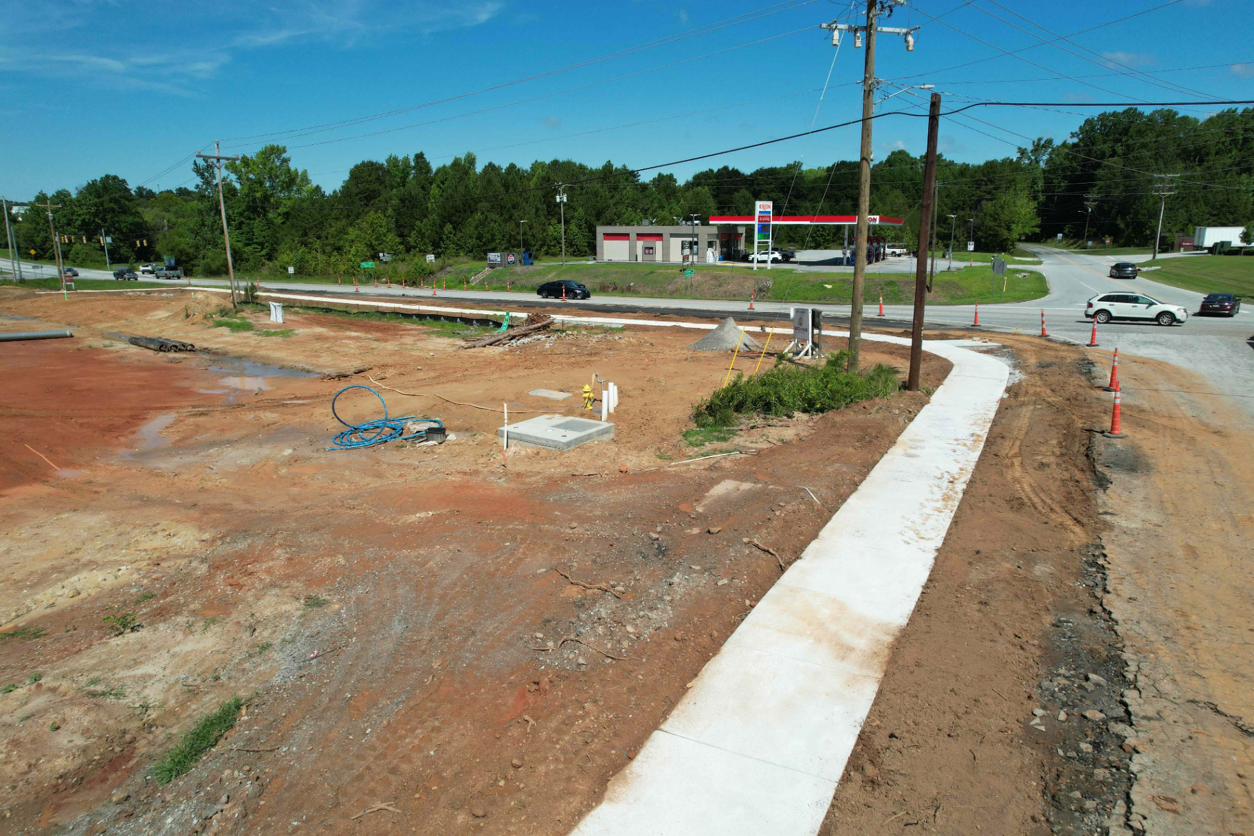 Aerial view of a new roadside sidewalk and curb ribbon running past a cleared commercial pad, with an Exxon station and highway intersection in the background.